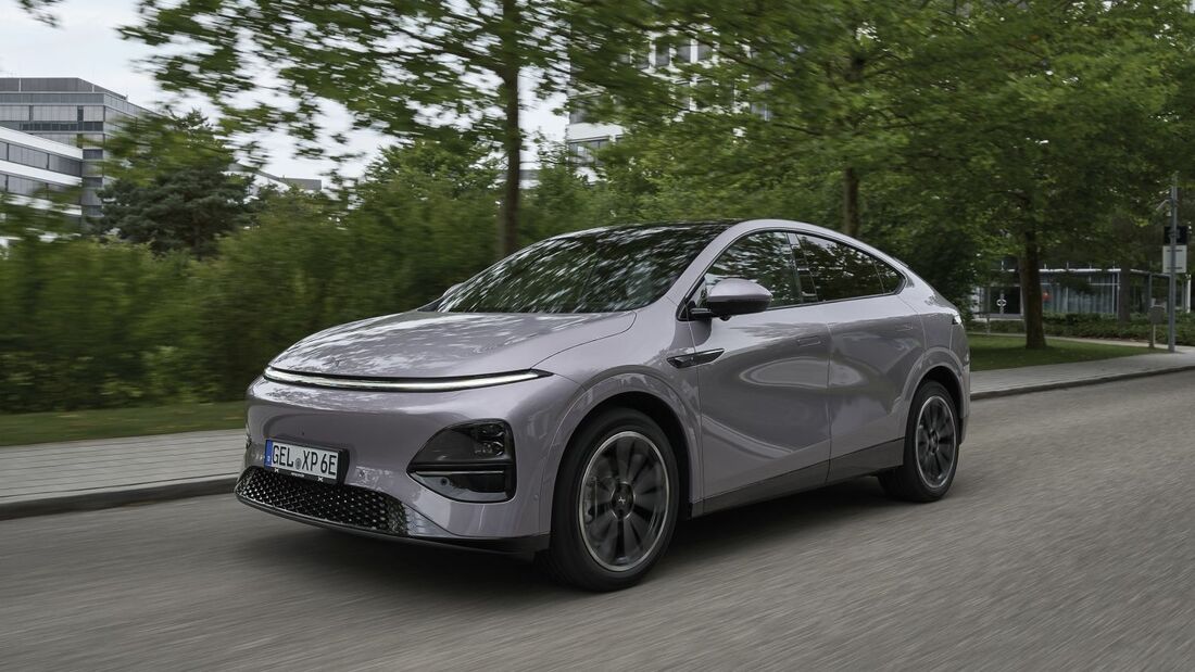 Xpeng G6 RWD Standard Range in Grau, Front-Seitenansicht während der Fahrt auf einer Straße mit Bäumen und Gebäuden im Hintergrund.