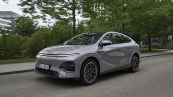 Xpeng G6 RWD Standard Range in Grau, Front-Seitenansicht während der Fahrt auf einer Straße mit Bäumen und Gebäuden im Hintergrund.