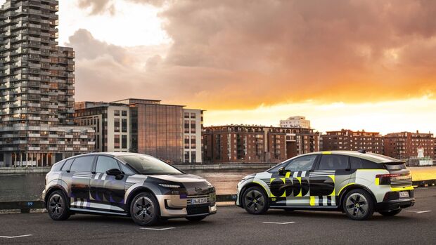 Zwei Audi A2 E-Tron Prototypen stehen mit unterschiedlicher Tarnbeklebung an einem Hafen vor moderner Stadtkulisse.