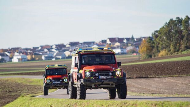 Zwei Feuerwehr-INEOS-Grenadier fahren hintereinander auf einer Landstraße mit Ortschaft im Hintergrund.