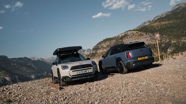 Zwei MINI Countryman Untamed Edition stehen auf einem Schotterplatz in einer Berglandschaft, links ein helles Modell mit Dachbox.