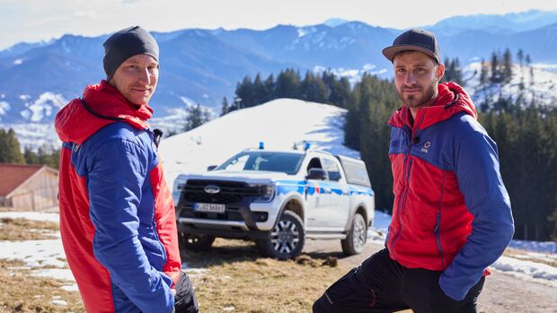 Zwei Mitglieder der Bergwacht Bayern stehen vor einem Ford Ranger Plug-in-Hybrid in verschneiter Alpenlandschaft.