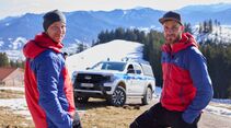 Zwei Mitglieder der Bergwacht Bayern stehen vor einem Ford Ranger Plug-in-Hybrid in verschneiter Alpenlandschaft.
