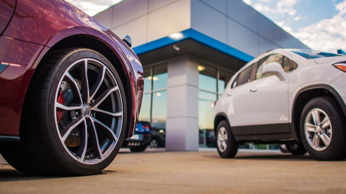 Zwei Neuwagen stehen vor einem Autohaus, im Vordergrund ein Fahrzeugrad, im Hintergrund ein weiteres Fahrzeug auf dem Händlergelände.