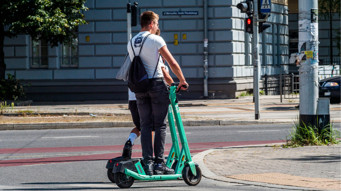 Zwei Personen fahren mit einem E-Scooter an einer Kreuzung in der Stadt. Eine Ampel zeigt Rot.