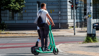 Zwei Personen fahren mit einem E-Scooter an einer Kreuzung in der Stadt. Eine Ampel zeigt Rot.