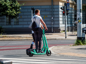 Zwei Personen fahren mit einem E-Scooter an einer Kreuzung in der Stadt. Eine Ampel zeigt Rot.
